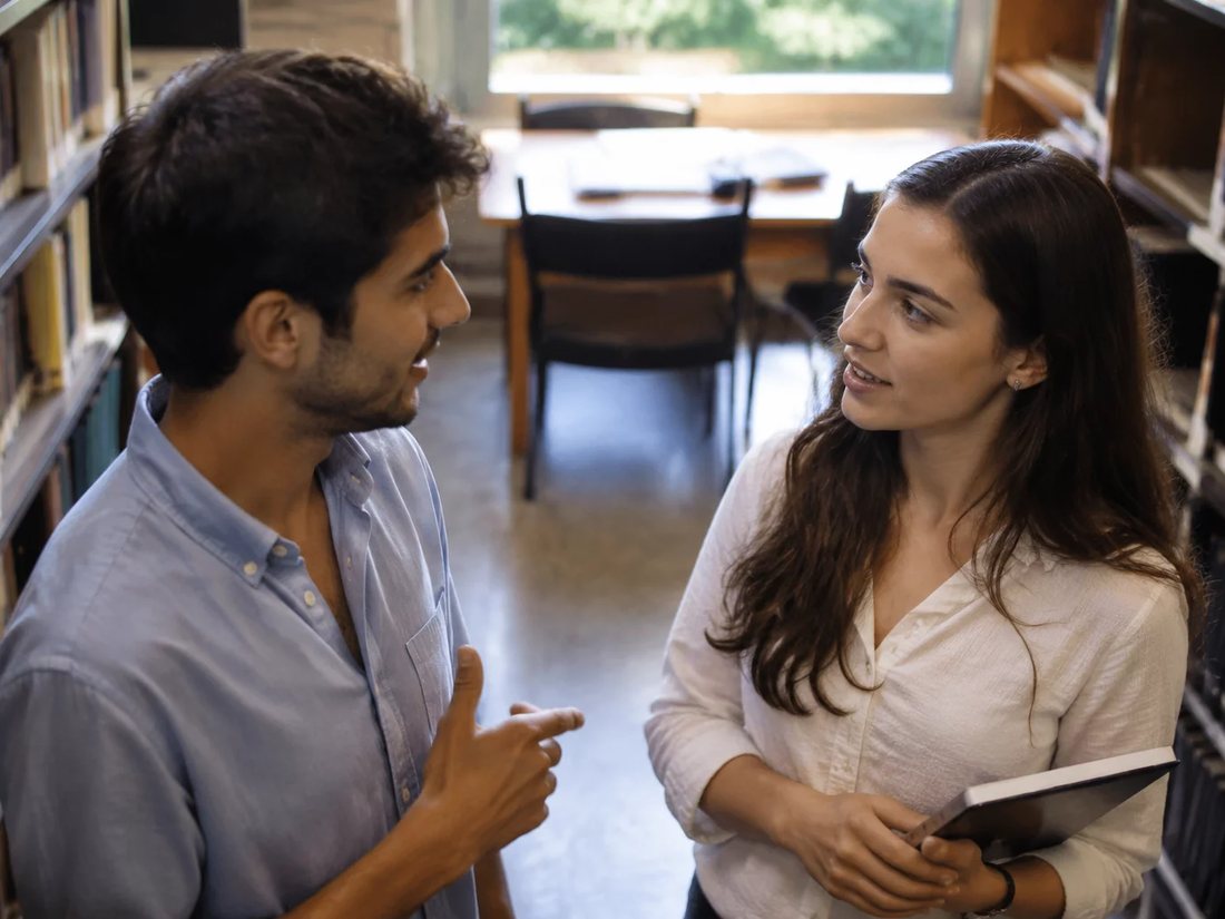 Conversa em pé entre as estantes da escola, com folhas de anotação na mão de quem discute